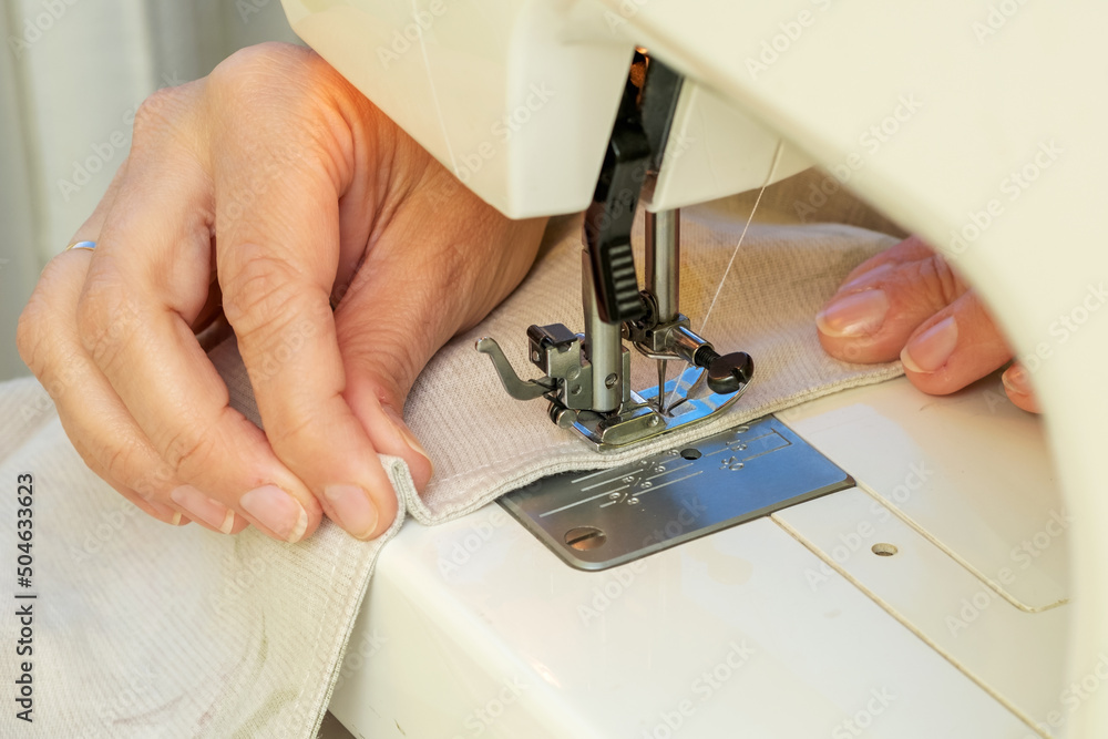 A woman sews on a sewing machine. Women's hands substitute the fabric for sewing under the foot of the sewing machine. - obrazy, fototapety, plakaty 