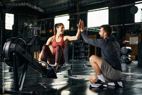 Happy athlete giving high-five to her personal trainer after exercising on rowing machine at gym Fototapeta