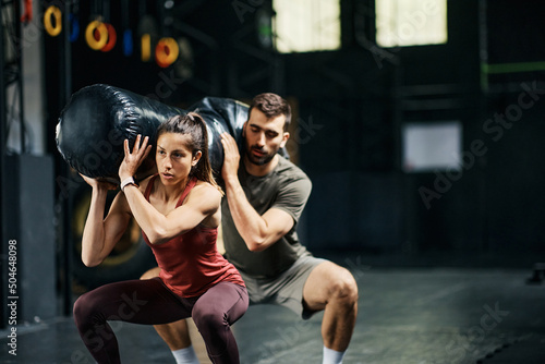 Athletic couple doing squats with sandbag on shoulders during cross training in gym. photo