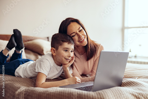 Pretty Caucasian mother and small teen son lying on bed and videochatting on laptop.