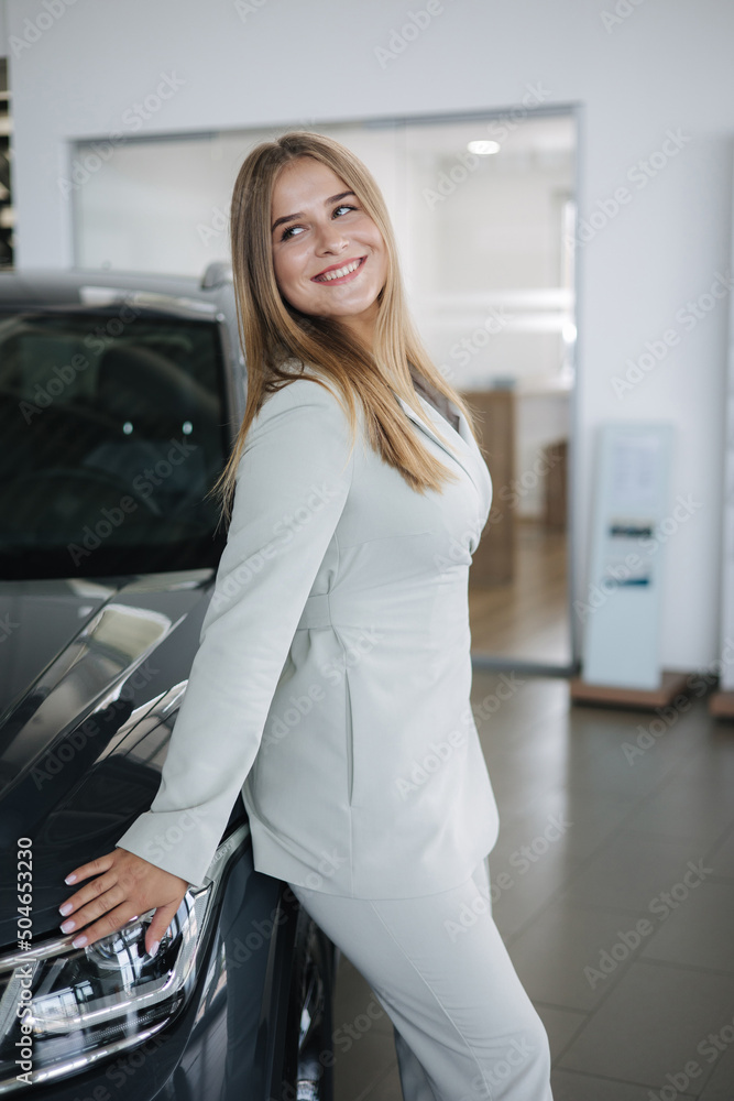 Portrait of gorgeous woman in showroom. Beautiful salesperson standing by car. Blond hair female after seles car Portrait of gorgeous woman in showroom. Beautiful salesperson standing by car. Blond hair female after seles car