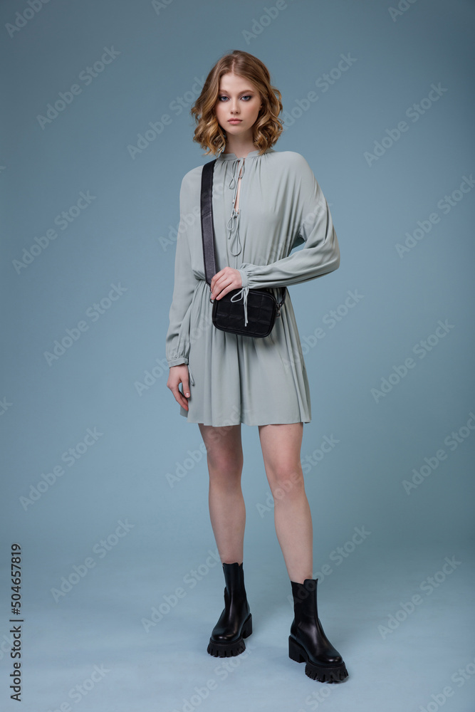 Fototapeta premium High fashion photo of a beautiful elegant young woman in pretty soft blue gray dress, boots, black handbag posing on blue background. Slim figure. Studio shot. Monochrome