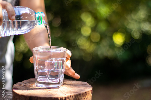 Hand holding drinking water bottle pouring water into glass on wooden table on blurred green nature background.