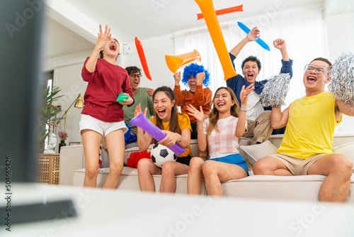 Fototapeta Naklejka Na Ścianę i Meble -  Group of Asian people friends sit on sofa watching and cheering soccer games competition on TV together at home. Happy man and woman sport fans shouting and celebrating sport team victory sports match