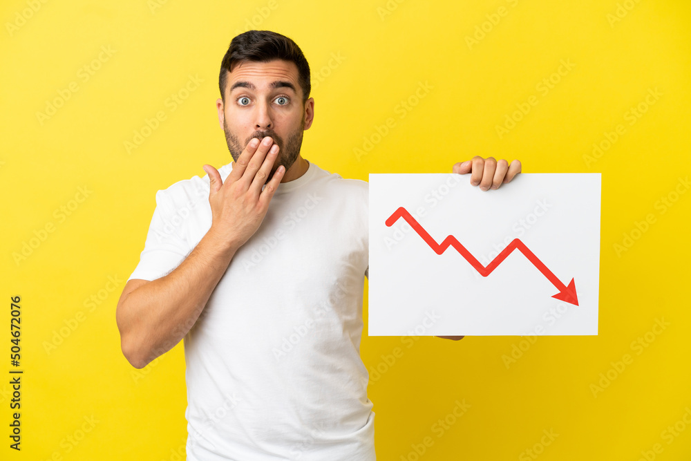 Young handsome caucasian man isolated on yellow background holding a sign with a decreasing statistics arrow symbol with surprised expression Young handsome caucasian man isolated on yellow background holding a sign with a decreasing statistics arrow symbol with surprised expression