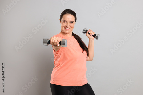 Obraz na plátně Happy overweight woman doing exercise with dumbbells on grey background