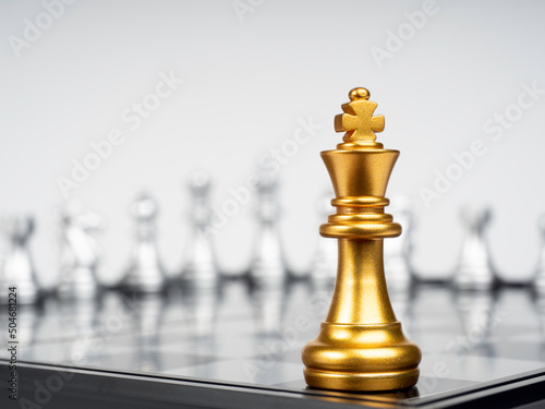 Fotografi The Golden king chess piece standing on chessboard corner in front of many silver chess pieces on white background