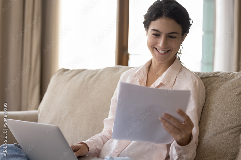 Smiling young woman learn banking paper notification about last mortgage payment while pay bills seated on sofa with laptop. Satisfied millennial female read great news in letter notice feels happy Smiling young woman learn banking paper notification about last mortgage payment while pay bills seated on sofa with laptop. Satisfied millennial female read great news in letter notice feels happy