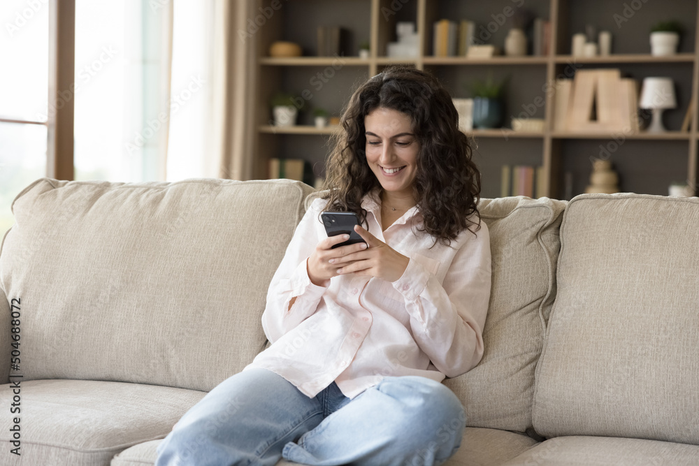 Attractive teenage girl sits on couch with smart phone. Young smiling woman spend time at home holding modern cellphone, share messages with friend, enjoy e-dating services, texting sms. Tech concept Attractive teenage girl sits on couch with smart phone. Young smiling woman spend time at home holding modern cellphone, share messages with friend, enjoy e-dating services, texting sms. Tech concept