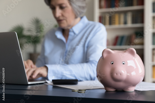 Toy pink piggy bank on work table of mature business woman using laptop, doing financial paperwork. Senior lady counting savings, taxes. Finance management, insurance, personal budget concept