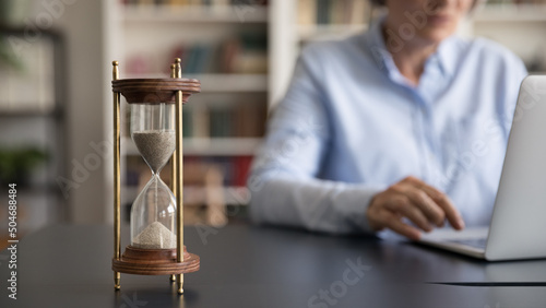 Vintage sand watch on work table of senior business professional lady using laptop computer on blurred background. Hourglass on workplace desk, closed up object. Business time management concept