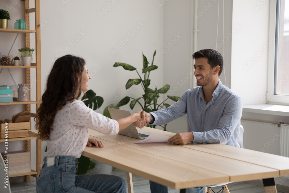 Happy young business leader man employing worker, shaking woman hand over work table after successful job interview, hiring professional, holding paper resume, laughing, smiling Happy young business leader man employing worker, shaking woman hand over work table after successful job interview, hiring professional, holding paper resume, laughing, smiling