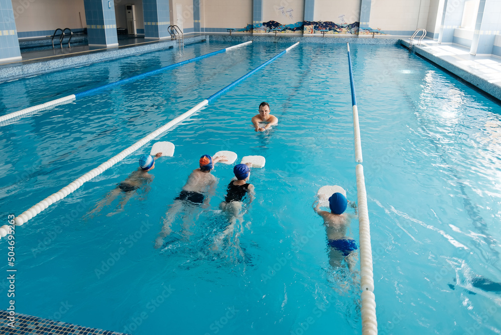 A group of boys and girls train and learn to swim in a modern swimming pool with an instructor. Development of children's sports. Healthy parenting and promotion of children's sports.