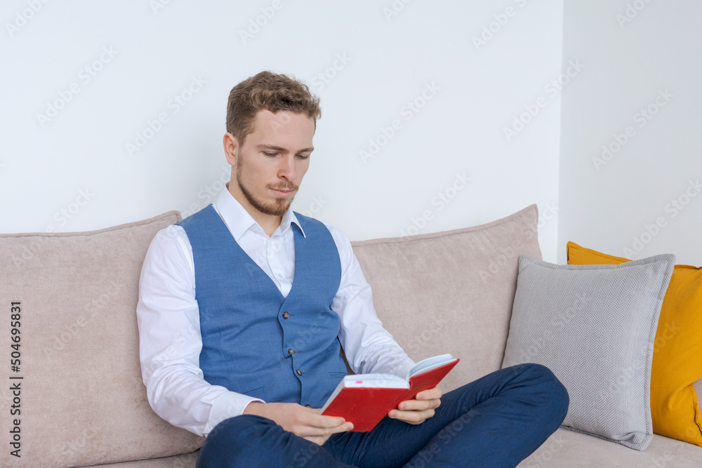 Focused young business man sits on couch with notebook in his hand and reads and analyzes an action plan for development at work in a blue suit. Caucasian businessman in the office on the couch Focused young business man sits on couch with notebook in his hand and reads and analyzes an action plan for development at work in a blue suit. Caucasian businessman in the office on the couch