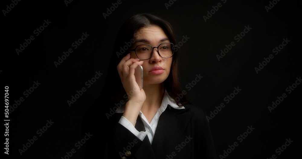 Business woman with glasses and long brown hair stands on a black background in office clothes,answers a phone call, talks on a smartphone with a frustrated face, sighs sadly Business woman with glasses and long brown hair stands on a black background in office clothes,answers a phone call, talks on a smartphone with a frustrated face, sighs sadly