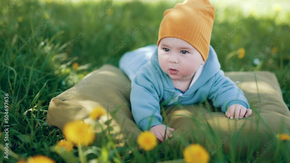 Lovely baby in blue overall and orange cap lies on his belly. Boy resting on the pillow in the green grass. Lovely baby in blue overall and orange cap lies on his belly. Boy resting on the pillow in the green grass.