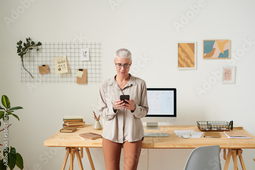 Content middle-aged businesswoman with blond hair standing at desk and checking messengers on smartphone in home office