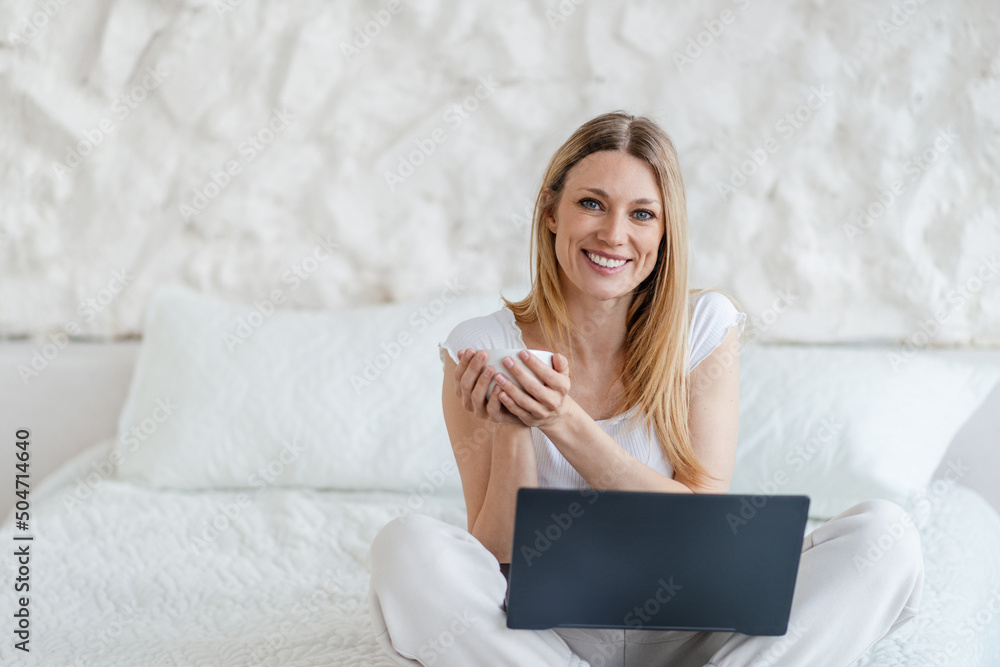 Smiling attractive young blond european woman with cup and laptop sits on bed in white bedroom Smiling attractive young blond european woman with cup and laptop sits on bed in white bedroom