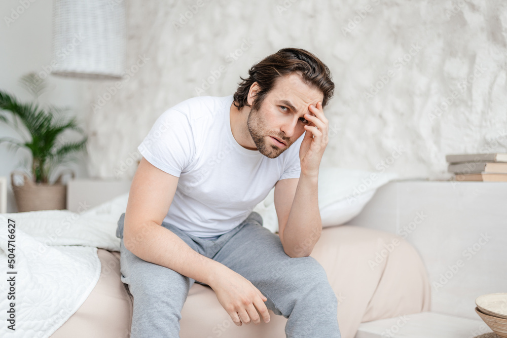 Sad millennial caucasian man with stubble sits on white bed, presses his hands to head and suffers from migraine Sad millennial caucasian man with stubble sits on white bed, presses his hands to head and suffers from migraine