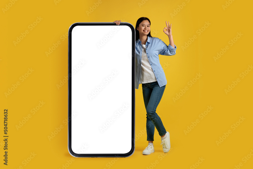Satisfied young chinese woman in casual show ok sign with hand, stands near huge phone with empty screen - obrazy, fototapety, plakaty 