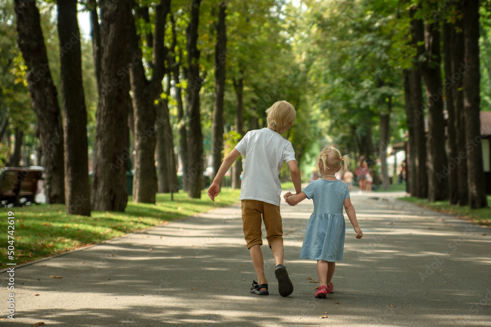 Little brother and sister walk in park. Older brother holds his sister's hand and walks along
