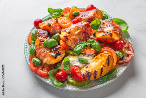 Delicious summer salad with grilled peaches, tomatoes, prosciutto and cheese in a plate on white table. Close up