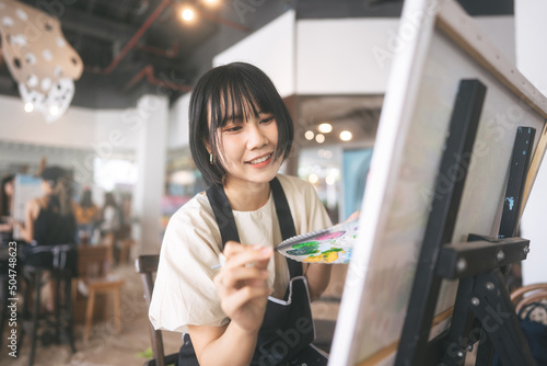 Fototapeta Naklejka Na Ścianę i Meble -  Portrait of happy young adult asian woman painting brush on canvas at workshop art lesson class.