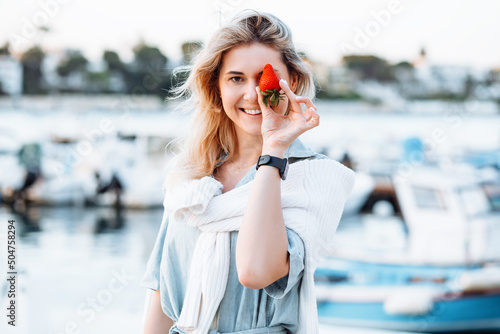 Foto Portrait of young smiling woman holding in hand ripe tasty strawberry on blurred background, walking along embankment with boats and yachts in resort city