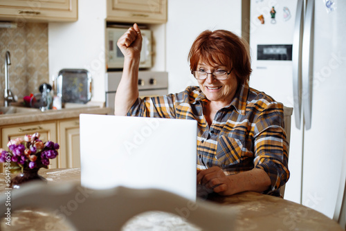 Happy mature lady looking at laptop screen sitting at table in kitchen. Advanced pensioner practicing computer skills and learning modern technology. Online communication and distant future concept