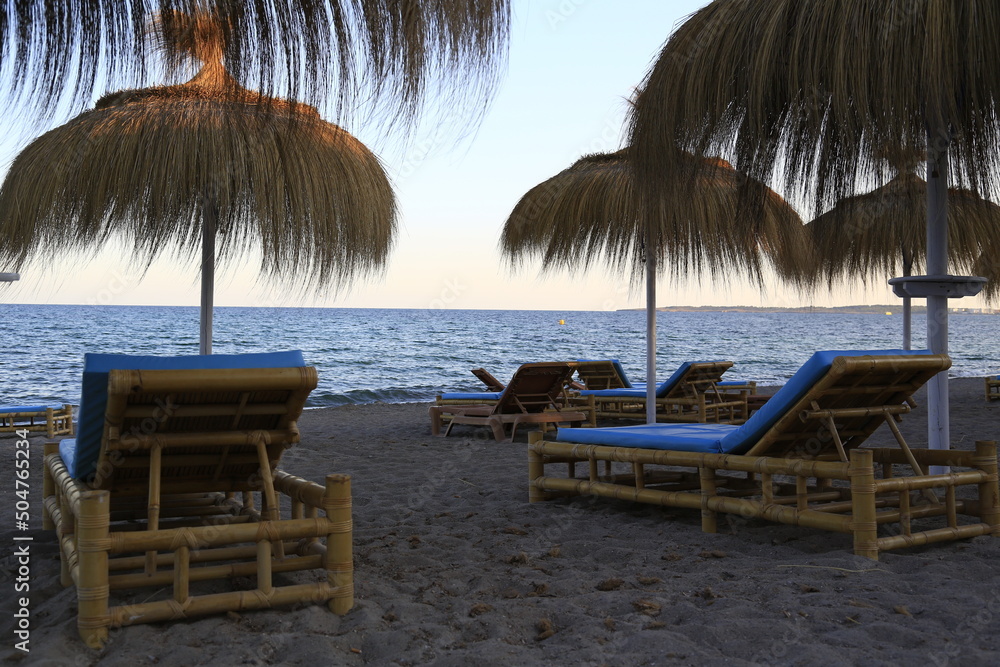 Empty comfortable rattan sunbeds under parasols made of seaweed on a sandy beach inviting to enjoy the view to the blue Mediterranean at sunset and evening atmosphere on Mallorca island, Spain