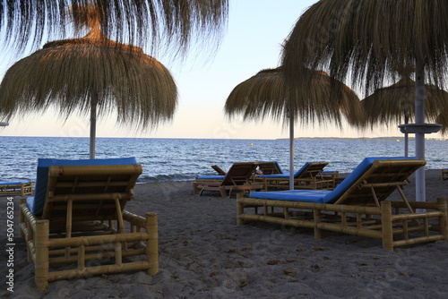 Empty comfortable rattan sunbeds under parasols made of seaweed on a sandy beach inviting to enjoy the view to the blue Mediterranean at sunset and evening atmosphere on Mallorca island  Spain