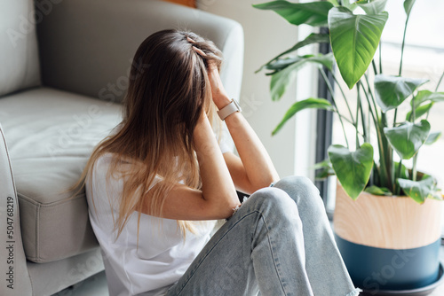 Unhappy young caucasian woman with blonde hair thinking about bad relationships problems, break up with boyfriend. Worried millennial girl sitting on floor in bedroom near chair and green plant alone photo