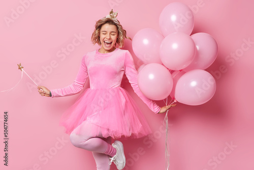 Upbeat cheerful female princess wears festive dress dances carefree holds magic wand and bunch of inflated helium balloons has fun on party isolated over pink studio background. Monochrome shot