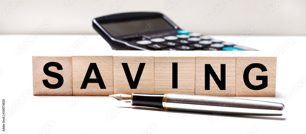 Obraz premium The light table has a black calculator, a fountain pen, and wooden cubes with the text SAVING. Business concept