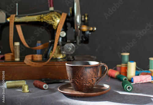 A clay mug with coffee on the background of an old vintage sewing machine and sewing accessories in the rays of the setting sun. Coffee time, work break, eco-friendly dishes.