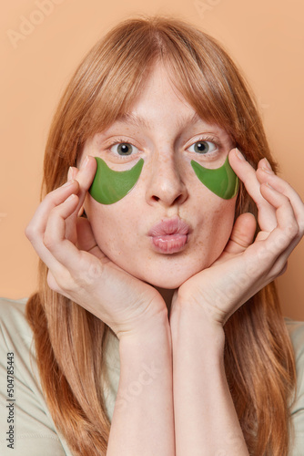 Wallpaper Mural Vertical shot of ginger freckled girl applies green collagen patches under eyes keeps lips folded looks directly at camera undergoes beauty procedures isolated over beige background