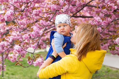 Young beautiful mother holds in her arms a little cute son. Mom and little baby boy in a flowering garden. Sakura.
