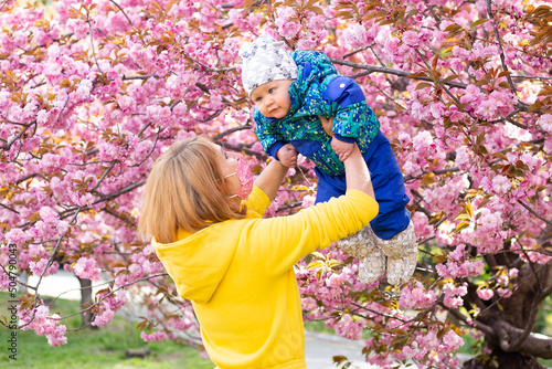 Young woman in medical mask with a child in her arms happy. Sakura blossom in spring Park. Joy of motherhood and child development. Cherry blossom tree in late spring