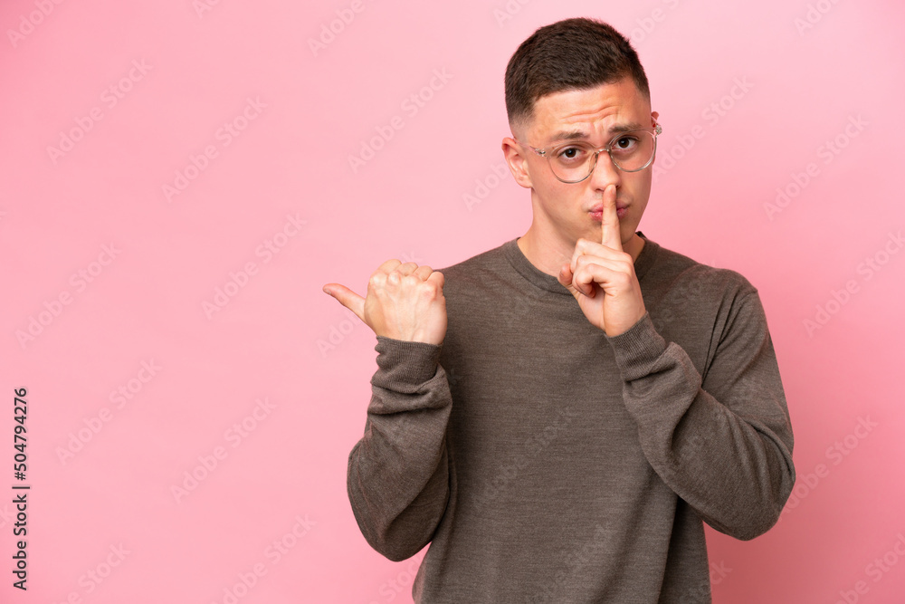 Young Brazilian man isolated on pink background pointing to the side and doing silence gesture Young Brazilian man isolated on pink background pointing to the side and doing silence gesture