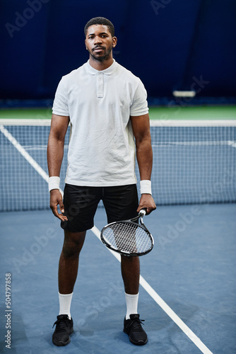 Full length portrait of African American tennis player looking at camera while posing confidently with racket at indoor court