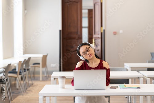 Fotografie, Tablou Tired Asian student girl in headphones doing gymnastics while listening webinar