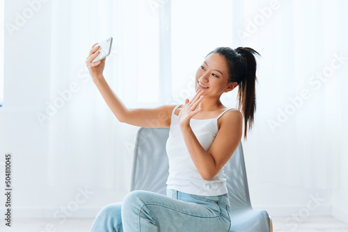 Tablou canvas Cheerful friendly tanned lovely young Asian woman greeting friend waving hand doing selfie video call at home interior living room