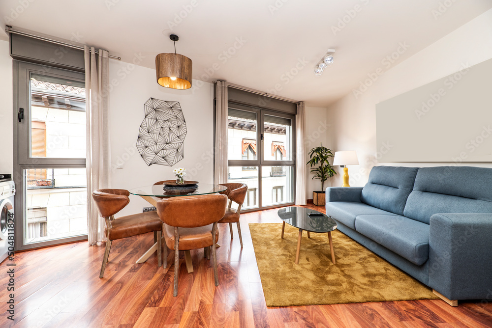 Fototapeta premium Living room with circular glass dining table with leather and wood chairs, reddish parquet floors and three-seater sofa bed upholstered in blue fabric, auxiliary coffee table on gold carpet