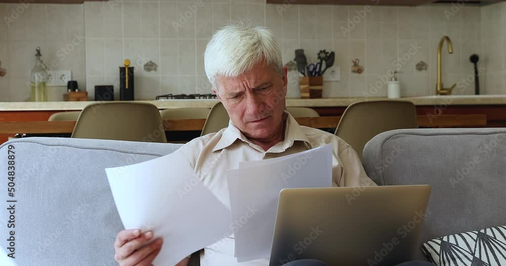 Stressed older male manage papers at home using laptop unable to pay bills deal with wrong work complicated interface of web bank app. Worried aged grandpa find loan debt reading financial documents