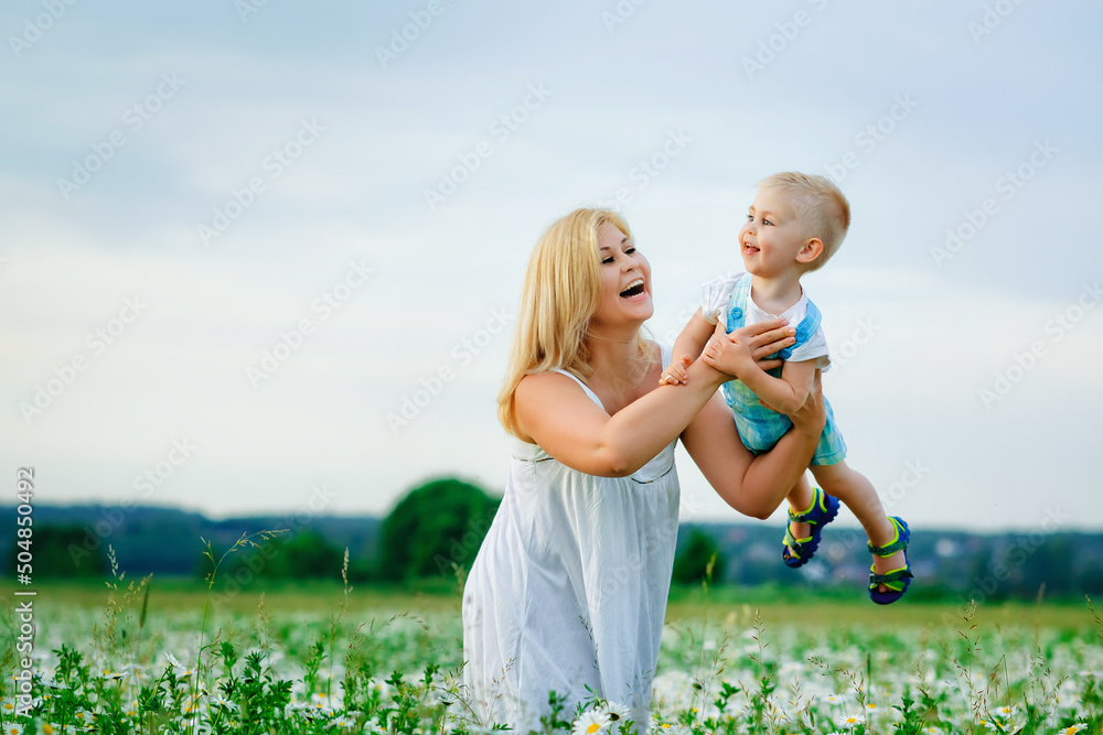 Fototapeta premium A plump woman and a small one-year-old boy in a field of daisies. A mother holds her son in her arms in a blooming field of camomile.