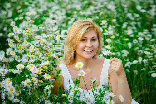 Portrait of a young, plump, beautiful woman resting on a chamomile field at sunset. A plus-size woman in a white sundress sits in a meadow with daisies.