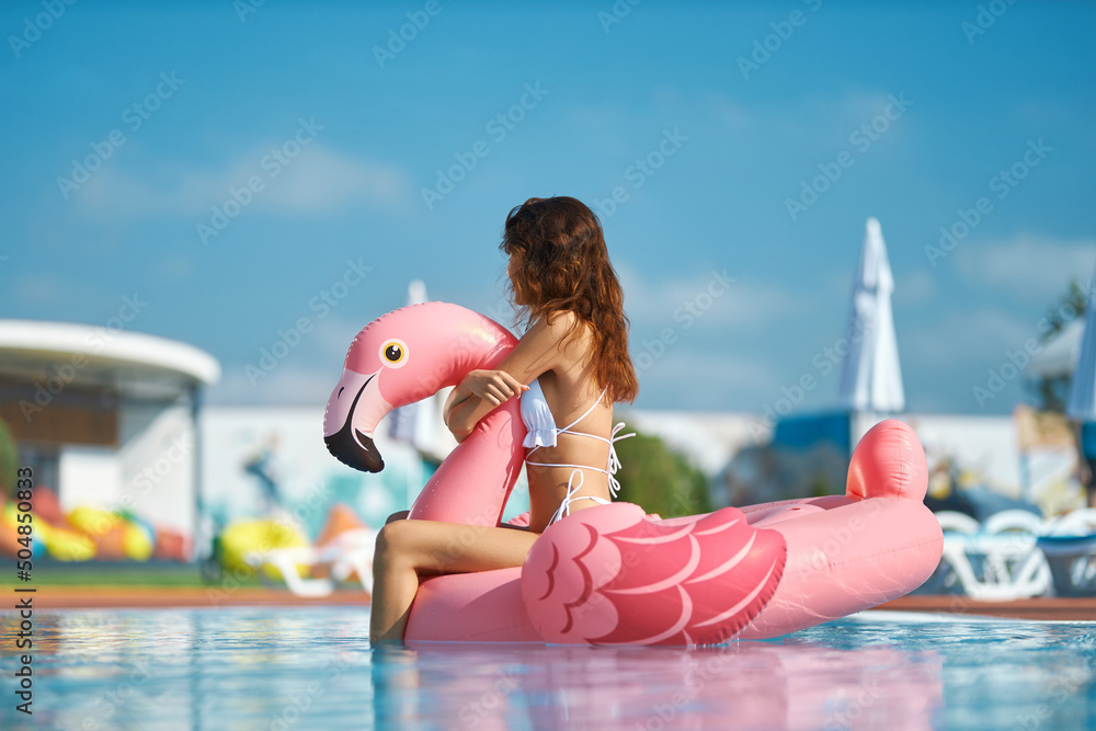Dreamy slim female sitting on float in pool with clean blue water at resort. Side view of long haired girl floating on pink inflatable flamingo in pool on sunny summer day. Concept of entertainment.  - obrazy, fototapety, plakaty 