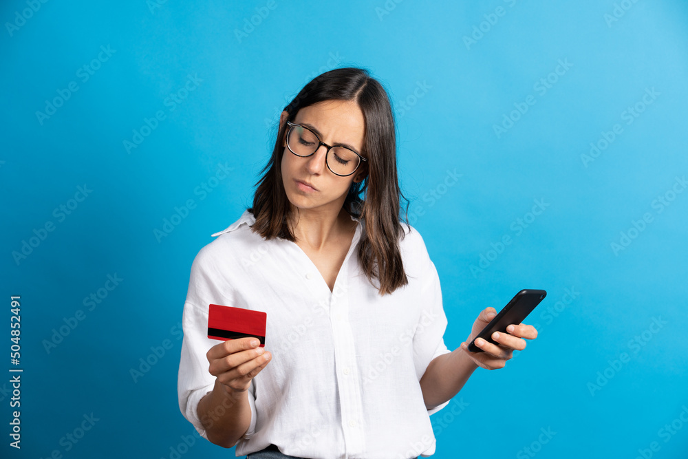 Serious pretty hispanic woman holding phone and credit card isolated on blue background. Electronic commerce concept
