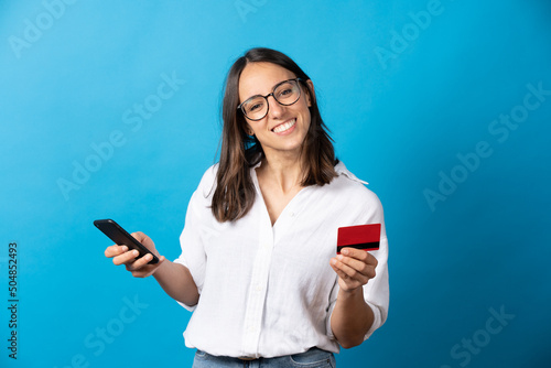 Fotografering Smiling hispanic woman shopping online and looking at camera isolated on blue background