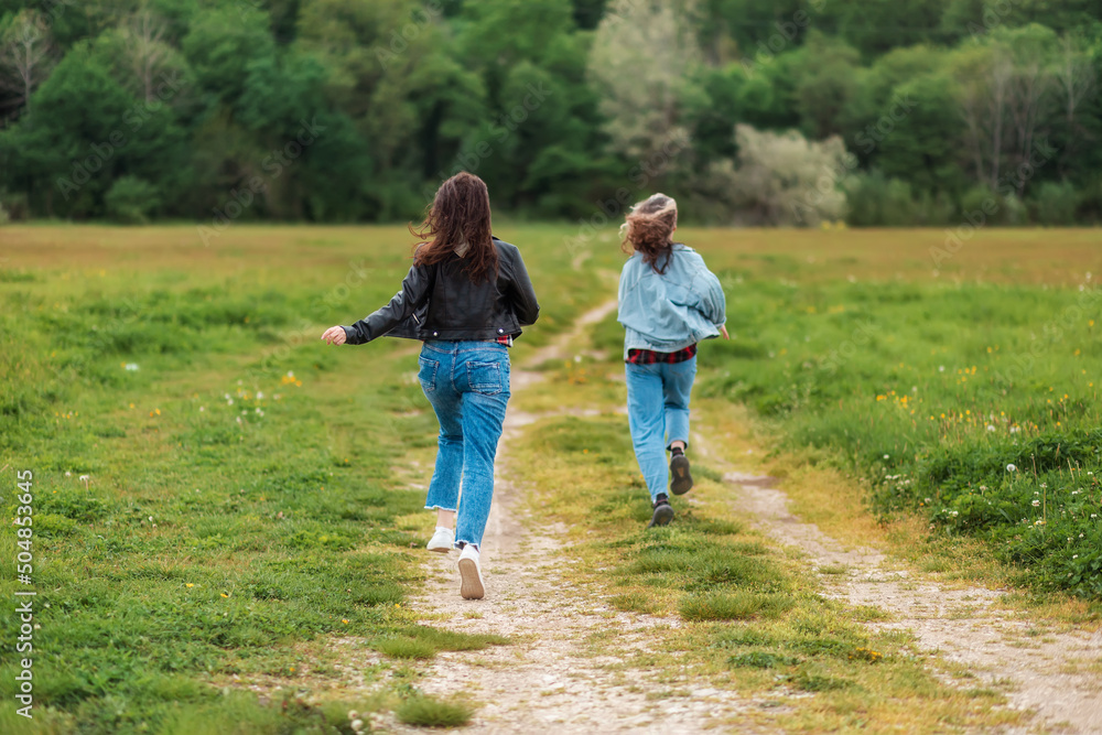 Family activity. A woman and a teen girl are joyfully running along a country road. Rear view. There is a forest in the background. The concept of freedom and happiness